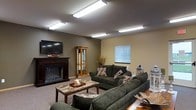 This single-floor space offers a warm and inviting atmosphere centered around a well-appointed lounge area. The room is anchored by a classic fireplace with a dark mantle, above which a wall-mounted flat-screen TV is positioned for easy viewing. Plush, dark green sofas and accent pillows are arranged around a stone-top coffee table, creating a comfortable and functional seating area suited for both relaxation and socializing. A wooden curio cabinet adds a traditional touch, while a decorative floral arrangement brings a subtle natural element to the space. The room is finished with neutral, earth-toned walls that contribute to its cozy ambiance. Bright flush-mount fluorescent lighting fixtures across the ceiling ensure the space is well-lit throughout. A window fitted with horizontal blinds allows for adjustable natural light. The carpeted flooring adds to the overall warmth and comfort of the environment. This thoughtfully arranged space balances practicality with a welcoming aesthetic, making it well-suited for communal use or leisure activities.