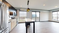 This single-floor unit showcases a contemporary industrial-inspired design, blending modern finishes with urban architectural details. The open-concept kitchen and living area serves as the heart of the space, anchored by a central island with a white quartz countertop beneath stylish pendant lighting. The kitchen is well-appointed with stainless steel appliances, a subway tile backsplash, two-tone cabinetry featuring light wood lower cabinets and dark upper cabinets, and durable light wood-tone flooring. Exposed metallic ductwork running along the ceiling adds an industrial character to the space.

Large, grid-paned windows throughout flood the interior with natural light and offer elevated views of the surrounding cityscape. A sliding glass door provides direct access to a private balcony, extending the living area outdoors. The bedrooms feature plush neutral carpet, ceiling fans, and generously sized windows with urban views.

A dedicated hallway connects the rooms efficiently, with dark-framed doors providing a consistent design accent. The bathroom features a floating wood-tone vanity, white quartz countertop, black matte fixtures, and a large framed mirror, maintaining the space's clean, modern aesthetic throughout.