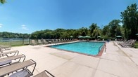 Located at 209 New Chalet Drive in Mohegan Lake, NY, this outdoor pool facility offers a well-maintained recreational environment surrounded by lush, mature trees. The centerpiece is a generously sized rectangular swimming pool with clear turquoise water, framed by a spacious concrete deck. The deck is furnished with rows of lounge chairs and teal patio umbrellas, providing ample space for sunbathing and relaxation. The pool is enclosed by a black metal safety fence, ensuring a secure environment, and is equipped with safety equipment including life rings. The backdrop of dense green foliage and open blue skies enhances the natural, refreshing ambiance of the space.

Adjacent to the pool area, the facility includes clean, accessible restroom facilities finished with neutral-toned tile flooring throughout. The restroom features a rich dark wood vanity with an integrated sink and a large wall mirror that reflects natural light admitted through a blinds-fitted window. The toilet area is equipped with stainless steel grab bars for accessibility. Wall-mounted paper towel and soap dispensers provide practical, hands-free convenience. The restroom's understated décor, including simple decorative accents, contributes to a tidy and functional space that complements the overall recreational facility.