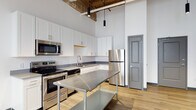 Located at 366 E Broad St in Columbus, Ohio, this single-floor space masterfully blends industrial character with modern finishes. The design is defined by striking exposed wood beam ceilings with visible ductwork and piping running overhead, creating a raw, loft-like aesthetic that contrasts beautifully with the clean white walls and light hardwood flooring throughout the main areas.

The kitchen is a standout feature, outfitted with stainless steel appliances including a range, microwave, and refrigerator, complemented by crisp white cabinetry with dark countertops. A large stainless steel worktable at the center adds both function and an industrial edge. Large windows with roller shades allow natural light to fill the space while maintaining privacy.

The open main area boasts soaring ceilings with pendant lighting and provides a versatile, airy environment. Additional private rooms are carpeted and brightened by natural light through individual windows, offering quieter, more enclosed settings. The bathroom is clean and modern, featuring a white vanity, tile flooring, and a mirror with overhead lighting.

Throughout, the combination of exposed structural elements, contemporary fixtures, and thoughtful layout makes this a distinctive and highly functional space suited for a variety of uses.