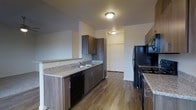 This single-floor residence offers a thoughtfully designed layout centered around a sleek, modern kitchen. The kitchen is outfitted with rich, dark wood cabinetry that provides ample storage space, both above and below the countertops. Granite-style countertops run along the perimeter, offering generous prep space, and a double-basin sink is conveniently positioned along the lower counter run. Black appliances, including a refrigerator, stove, dishwasher, and an over-range microwave, lend a cohesive, contemporary aesthetic throughout the space. The kitchen flows seamlessly across warm hardwood-style flooring that extends into the adjoining areas, adding a sense of continuity and warmth. Flush-mount ceiling lighting provides even, functional illumination throughout. Just beyond the kitchen, a hallway leads to additional living areas, including what appears to be a spacious closet with bi-fold doors, hinting at generous storage options throughout the unit. The neutral wall tones and clean lines of the interior create a bright, welcoming atmosphere that complements the modern finishes. Overall, the space combines practical functionality with a contemporary design sensibility, making it an inviting and efficient living environment.