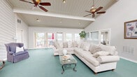 Located at 65 Villa Rd in Greenville, SC, this single-floor space is centered around a generously sized living room that makes a strong architectural impression. The room features dramatic vaulted ceilings with exposed white-painted beams, a ceiling fan, and recessed lighting that together create a bright and airy atmosphere. Soft teal carpeting runs throughout, adding a warm, inviting tone to the open layout.

A white shiplap-paneled bar counter with granite-style surfaces and barstool seating defines one end of the space, adjacent to a kitchen area with white cabinetry — ideal for entertaining. Tall ceilings with clerestory windows on one side allow abundant natural light to pour in, enhancing the sense of volume and openness.

The living area is generously proportioned, comfortably accommodating a large sectional sofa, accent chairs, and a glass coffee table. French doors lead directly to an outdoor deck, offering views of the surrounding neighborhood and bringing the outdoors in. Additional doors provide access to what appears to be a fitness area, adding functional versatility to the space.

Decorative wall art and potted greenery contribute to the welcoming ambiance, while the crisp white walls and beamed ceiling maintain a clean, cohesive aesthetic throughout.