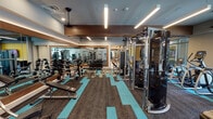 Located at 1133–1147 College Ave in Fort Worth, Texas, this well-appointed fitness center is designed to accommodate a wide range of workout needs across a single, thoughtfully organized floor. The space is divided into distinct functional zones, each outfitted with professional-grade equipment.

The main strength training area features an extensive dumbbell rack, multiple weight benches, cable machines, power racks, and a full-length mirrored wall that enhances the sense of openness. Stylized teal and charcoal patterned carpet tiles define the workout zones, adding a modern visual identity to the floor. Overhead, linear LED lighting and ceiling fans provide consistent illumination and airflow throughout.

The cardio zone is lined with floor-to-ceiling windows that frame outdoor views, flooding the area with natural light. Matrix-brand treadmills, ellipticals, and stair climbers are arranged to allow users to exercise while enjoying the exterior scenery.

A dedicated group fitness studio offers a clean, open layout with light wood-toned flooring, full mirrored walls, and a large wall-mounted display screen. The studio is stocked with stability balls, resistance bands, foam rollers, and TRX-style suspension trainers, making it well-suited for classes or individual functional training sessions.

A transitional lounge area with a mounted TV, cubbies, and large windows provides a welcoming entry point to the facility. The overall design balances energetic aesthetics with a functional, professional fitness environment.