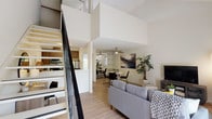 This two-story residence is designed with a functional, open layout that maximizes the sense of space throughout. The main living area is anchored by soaring double-height ceilings, which lend an airy, expansive quality to the room. Light wood-style flooring runs through the open-plan living and dining zones, unifying the spaces seamlessly. A ceiling fan overhead adds a practical touch, while a built-in shelving unit provides display and storage options. The living area opens toward a private balcony, allowing natural light to filter in and offering views of surrounding greenery.

The galley-style kitchen features warm wood cabinetry, light countertops, stainless steel appliances including a refrigerator, range, and dishwasher, and a single-basin sink. A staircase with dark metal railings leads to the upper level, where the bedrooms are located. The carpeted bedrooms offer a quieter, cozier atmosphere, with angled ceilings adding architectural character. The second bedroom is versatile enough to accommodate multiple sleeping arrangements. A bathroom on the upper level includes a vanity with wood cabinetry and a well-lit mirror. A mirrored sliding-door closet provides generous storage space adjacent to the bathroom.