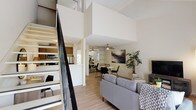This two-story residence is designed with a functional, open layout that maximizes the sense of space throughout. The main living area is anchored by soaring double-height ceilings, which lend an airy, expansive quality to the room. Light wood-style flooring runs through the open-plan living and dining zones, unifying the spaces seamlessly. A ceiling fan overhead adds a practical touch, while a built-in shelving unit provides display and storage options. The living area opens toward a private balcony, allowing natural light to filter in and offering views of surrounding greenery.

The galley-style kitchen features warm wood cabinetry, light countertops, stainless steel appliances including a refrigerator, range, and dishwasher, and a single-basin sink. A staircase with dark metal railings leads to the upper level, where the bedrooms are located. The carpeted bedrooms offer a quieter, cozier atmosphere, with angled ceilings adding architectural character. The second bedroom is versatile enough to accommodate multiple sleeping arrangements. A bathroom on the upper level includes a vanity with wood cabinetry and a well-lit mirror. A mirrored sliding-door closet provides generous storage space adjacent to the bathroom.