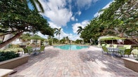 This Olen Living community amenity space offers a well-appointed outdoor retreat with a distinctly Florida-inspired character. At its centerpiece is a generously sized resort-style swimming pool, framed by a broad, paver-laid pool deck furnished with lounge chairs and shaded by green market umbrellas. Towering palm trees and mature tropical foliage surround the pool area, creating a lush, private atmosphere beneath open blue skies.

Adjacent to the pool, manicured garden beds feature raised planter walls with vibrant flowering accents, including eye-catching red ceramic urns bursting with color. Winding paver pathways connect the various outdoor zones, linking the pool deck to quieter garden seating areas ideal for relaxation.

The covered entrance portico is a standout architectural feature, boasting soaring double-height ceilings with ceiling fans, classic colonial-style columns, and decorative moldings. Elegant glass-paneled double doors open to the interior, allowing views directly through to the shimmering pool beyond. Outdoor seating with a round table and green chairs makes this covered area a comfortable transitional space between indoors and out.

The building's exterior presents a refined stucco façade with detailed millwork, well-integrated into the surrounding tropical landscaping, creating a cohesive and welcoming community environment throughout.