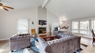 Located in Fremont, CA (94538), this single-floor communal space offers a thoughtfully designed layout encompassing a living room, dining room, and kitchen. Vaulted ceilings with exposed beams and ceiling fans lend an open, airy feel throughout, while warm wood-look flooring runs seamlessly across the space. The living room serves as a central gathering area, anchored by a tile-surround fireplace with a mounted flat-screen TV above it. Plush tufted gray sofas arranged around a wooden coffee table on a blue area rug create an inviting lounge atmosphere. Large windows and sliding glass doors with vertical blinds flood the room with natural light and provide views of the surrounding neighborhood. The dining area accommodates a substantial wooden table with matching mission-style chairs, lit by a decorative pendant light above, and features a pass-through window connecting to the kitchen. The kitchenette is efficiently outfitted with white cabinetry, a compact stainless steel refrigerator, microwave, drip coffee maker, water dispenser, and a sink — ideal for light food and beverage service. The overall ambiance is clean, bright, and welcoming, making this a functional and comfortable shared space.