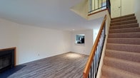 This well-designed multi-level residence spans three floors, offering a thoughtfully organized layout with functional living spaces throughout. The main living area features smooth hardwood-style laminate flooring in a warm gray tone, clean white walls, and recessed lighting that creates a bright, welcoming atmosphere. A pass-through window connects the living area to the kitchen, which is equipped with cabinetry and a full-size refrigerator — a practical detail that promotes an open, connected feel between spaces. A staircase with a warm wood handrail and dark metal balusters anchors the main floor, providing access to the upper levels. Carpeted steps add comfort underfoot while complementing the overall design. The upper level features a railed balcony overlook, adding a sense of vertical openness and architectural depth to the interior. The clean, neutral color palette throughout ensures versatility and timeless appeal. With three floors of living space, this home offers ample room for a variety of living arrangements, making it well-suited for those seeking a structured, multi-level environment with modern finishes and an open, airy feel.