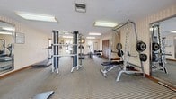 This fitness center offers a thorough selection of exercise equipment arranged across a generously sized, single-level space. The room is finished with neutral striped wallcovering, a textured ceiling, and a grid-patterned carpet that provides both durability and comfort underfoot. Fluorescent lighting is evenly distributed throughout, ensuring the entire area is well-illuminated during workouts.

The cardio section includes multiple treadmills, an elliptical trainer, and a rowing machine, providing a variety of options for aerobic exercise. The strength-training area is equally well-appointed, featuring a Magnum cable machine system, a power rack loaded with weight plates, a dedicated dumbbell rack with a full range of free weights, and an adjustable workout bench. A large wall-mounted mirror enhances the sense of space and allows users to monitor their form.

Natural light filters in through several windows along one wall, offering views of outdoor greenery and contributing to an open, inviting atmosphere. Storage is provided via a set of louvered wooden closet doors along one wall. A hallway connects the fitness center to adjacent facilities, adding to the overall convenience of the space.