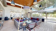Located at 3900 W Northwest Hwy in Dallas, TX, this striking single-floor community lounge showcases a bold contemporary design that seamlessly blends architectural drama with curated interior details. The space is anchored by soaring vaulted ceilings clad in warm wood paneling, creating a rich contrast against the clean white walls and light hardwood-style flooring below. Floor-to-ceiling windows and sliding glass doors line the exterior walls, flooding the interior with an abundance of natural light while offering sweeping views of the lush, tree-lined outdoor surroundings. The living area is generously appointed with large sectional sofas in shades of grey and purple, accented with vibrant red throw pillows, complemented by woven lounge chairs with striking red metal frames. Statement pendant lighting in muted blue-grey and black tones hangs overhead, adding to the layered visual appeal. A textured brick accent wall serves as a backdrop for a large wall-mounted flat-screen television, flanked by sculptural resin side tables. Original artwork is displayed throughout, including a bold abstract canvas and a striking mixed-media metal wall sculpture. A shuffleboard table adds a recreational focal point, while a bar-style counter area with decorative globe lighting rounds out the space. The overall ambiance is modern, vibrant, and inviting.