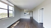 This single-floor residence showcases a clean, contemporary design centered around an open-concept living and kitchen layout. Rich, dark hardwood flooring flows throughout the space, providing a striking contrast against the light gray walls and crisp white trim. The expansive living area is flooded with natural light through large windows that frame views of lush greenery, creating a bright and welcoming atmosphere.

The kitchen is thoughtfully integrated into the living space via a peninsula-style countertop, offering both a functional workspace and a natural divider between the two areas. The kitchen features stainless steel appliances, including a refrigerator and microwave, alongside modern gray cabinetry that extends to the upper portion of the walls. Two stylish pendant lights hang above the kitchen area, adding a warm, focused glow that complements the overall aesthetic.

The open sightlines from the living area through to the kitchen and into the hallway beyond give the space a continuous, flowing feel. The clean lines, neutral color palette, and contemporary finishes throughout contribute to a polished and versatile interior that is both functional and visually appealing. The single-floor layout ensures ease of movement and accessibility across the entire space.
