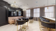 This refined single-floor space exudes understated glamour through a carefully curated blend of Art Deco and contemporary design. The main area features light-toned herringbone hardwood flooring that contrasts elegantly with the deep charcoal and black wall treatments, creating a dramatic yet inviting atmosphere. Multiple crystal and brass chandeliers cascade from the white ceiling, casting a warm, luminous glow throughout the space.

The lounge and dining area is furnished with black upholstered seating on brushed gold bases, black marble-topped high tables, and a plush blush-toned banquette running along one wall. A large mirror panel amplifies the sense of depth and light. A floral-patterned accent chair adds a bold, artistic touch.

The adjoining kitchen is sleek and gallery-like, with floor-to-ceiling dark wood cabinetry and expansive white stone countertops lining both sides. Pendant lighting and framed artwork integrated into mirrored panels give the kitchen a distinctly editorial character.

The entrance features floor-to-ceiling glass doors etched with the venue's branding, flanked by dark panelled walls that set the tone for the sophisticated interior. Sheer taupe curtains dress the windows throughout, diffusing natural light softly across the entire space.