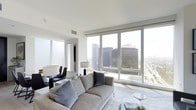 This sophisticated high-rise residence offers a refined urban living experience defined by clean lines, a neutral palette, and an abundance of natural light. The open living area is anchored by expansive floor-to-ceiling windows that frame striking panoramic views of the surrounding city skyline, including prominent downtown high-rises and the sprawling cityscape beyond — a defining architectural feature that brings the energy of the city indoors.

The living space is thoughtfully designed with a minimalist aesthetic, featuring smooth white ceilings, sleek wall finishes, and light-toned flooring that enhances the airy, open atmosphere. A comfortable seating area with a contemporary sofa and accent pillows is positioned to take full advantage of the sweeping views, complemented by a glass coffee table that adds to the modern sensibility. A dining area with upholstered chairs and a stylish lamp is visible in the background, offering a seamless transition between living and dining spaces.

Natural light floods the interior throughout the day, creating a bright and welcoming ambiance at every hour. The overall design is cohesive and understated, prioritizing space, light, and the dramatic visual connection to the urban environment outside. This single-floor residence delivers a compelling blend of contemporary design and elevated city living.