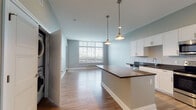 Located at 220 N Patterson Blvd in the heart of downtown Dayton, Ohio, this single-floor unit offers a well-designed open-concept layout with contemporary finishes throughout. The living area is bright and inviting, featuring light wood-style flooring, soft neutral wall tones, and a expansive bank of windows fitted with blinds that frame views of the Dayton skyline. A door leads directly to a private balcony, further connecting the interior to the urban surroundings.

The kitchen is a functional centerpiece of the space, showcasing white shaker-style cabinetry, dark countertops, and a full suite of stainless steel appliances including a range, microwave, dishwasher, and refrigerator. A central island with pendant lighting provides additional prep and seating space, creating a natural divide between the kitchen and living area.

The bedroom is carpeted for comfort and features a large window offering city views, complemented by neutral gray walls. The bathroom is well-appointed with a dual-sink vanity topped with dark counters, white cabinetry, a large mirror with overhead lighting, and a walk-in shower with sliding glass doors set over tile flooring. The overall design balances modern aesthetics with practical, comfortable living in a prime downtown location.