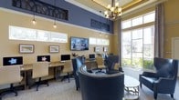 This single-story business center combines functionality with sophisticated design. The room features soaring ceilings accented by decorative arch-patterned transom panels in dark metal, creating a refined architectural detail against the two-tone walls of warm gold and deep navy blue. An elegant multi-light chandelier anchors the space from above, complemented by pendant lighting that adds warmth throughout.

Workstations with granite-topped desks line the walls, each equipped with desktop computers and ergonomic task chairs, providing ample workspace for productivity. A wall-mounted flat-screen TV serves as a central focal point between the workstations. Comfortable navy blue upholstered lounge chairs with nailhead trim offer an inviting seating area around a round side table, ideal for informal meetings or relaxed computing.

Large floor-to-ceiling windows along one wall flood the interior with natural light and frame lush outdoor greenery, creating a bright and airy atmosphere. Framed artwork adds a curated touch to the walls. The carpeted flooring and layered lighting contribute to a warm, professional ambiance well-suited for focused work or collaboration.