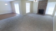 This single-story space centers around a generously sized living area finished with neutral, textured carpeting that extends throughout the main floor. A striking floor-to-ceiling stone fireplace anchors one corner of the room, serving as a prominent architectural focal point with its natural, rugged stonework and classic black firebox. The warm, beige-toned walls complement the earthy tones of the stone surround, creating a cohesive and inviting atmosphere.

Natural light fills the space through a combination of a large window fitted with white blinds and a set of French doors along the adjacent wall. The French doors offer direct access to the outdoors while allowing additional light to brighten the interior. A modest chandelier overhead adds a decorative touch to the living area.

The transition from carpet to hardwood or laminate flooring near the French doors subtly defines the entryway zone. The open layout of the living space provides flexibility for various furniture arrangements, making it well-suited for both everyday living and entertaining. Overall, the space conveys a comfortable, casual character with practical, durable finishes throughout.