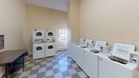 This shared laundry facility is a practical, well-equipped space designed to accommodate multiple users simultaneously. The room features a classic grey-and-white checkered vinyl tile floor that extends throughout, providing a clean and durable surface. Notably high ceilings lend an open, airy feel to the interior, with warm beige walls maintaining a neutral and functional aesthetic.

Along one wall, a row of commercial-grade top-load washing machines is neatly arranged, complemented by two stacked dryer units positioned nearby. A window allows natural light to filter into the laundry area, brightening the space during the day. A baseboard heater is also present, ensuring comfort during colder months.

A dedicated waiting area is thoughtfully incorporated into the layout, furnished with modern white molded chairs and a folding table, offering a convenient spot to sit while laundry cycles complete. Wall-mounted utility panels, a small storage cabinet, and posted notices—including no-smoking signage—contribute to the organized and regulated nature of the shared environment. The overall layout is efficient and straightforward, prioritizing functionality for everyday use.