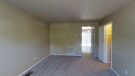 This two-floor residence offers a straightforward and functional layout with understated design elements throughout. The main living area is finished with wall-to-wall carpeting in a neutral gray tone, providing a comfortable and practical foundation for the space. The walls are painted in a warm beige hue, creating a simple, clean backdrop that lends itself well to a variety of décor styles.

A floor-level HVAC vent is integrated into the baseboard area, indicating a central heating and cooling system serving the space. The room benefits from natural light streaming in through what appears to be a sliding glass door, which offers a view of and access to an adjacent outdoor area. A ceiling fan is visible in the connected space beyond the doorway, suggesting additional climate control options.

The transition between rooms is defined by a standard doorway, hinting at a connected layout that flows between interior spaces. White baseboards and door trim provide a crisp contrast against the beige walls. The overall ambiance is modest and move-in ready, with a clean, unfurnished state that allows for flexible use of the available space across both floors.