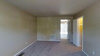 This two-floor residence offers a straightforward and functional layout with understated design elements throughout. The main living area is finished with wall-to-wall carpeting in a neutral gray tone, providing a comfortable and practical foundation for the space. The walls are painted in a warm beige hue, creating a simple, clean backdrop that lends itself well to a variety of décor styles.

A floor-level HVAC vent is integrated into the baseboard area, indicating a central heating and cooling system serving the space. The room benefits from natural light streaming in through what appears to be a sliding glass door, which offers a view of and access to an adjacent outdoor area. A ceiling fan is visible in the connected space beyond the doorway, suggesting additional climate control options.

The transition between rooms is defined by a standard doorway, hinting at a connected layout that flows between interior spaces. White baseboards and door trim provide a crisp contrast against the beige walls. The overall ambiance is modest and move-in ready, with a clean, unfurnished state that allows for flexible use of the available space across both floors.