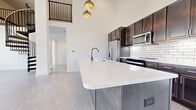 Located in the heart of Columbus, Ohio, this contemporary two-story home offers two bedrooms and four bathrooms across a generous, well-planned layout. The main floor commands attention with soaring double-height ceilings and a dramatic spiral staircase featuring sleek black metal railings and wood-toned steps, serving as a central architectural focal point. Large windows flood the open living area with natural light and offer urban views of the surrounding cityscape.

The kitchen is thoughtfully designed with rich dark cabinetry accented by gold hardware, a classic white subway tile backsplash, stainless steel appliances, and a large center island topped with bright white quartz countertops — ideal for both cooking and casual dining. Polished concrete-style flooring runs throughout the main living areas, lending an industrial-modern aesthetic.

The bedrooms are carpeted for warmth and comfort, with the well-sized primary bedroom featuring direct access to a walk-in closet outfitted with wire shelving systems. The four bathrooms are finished in a clean, minimal palette, including one bathroom with a fully tiled walk-in shower enclosed by black-framed glass doors. Additional spaces include a utility room, two closets, and multiple hallways. The upper level overlooks the main floor through black iron railings, reinforcing the home's cohesive modern design language.