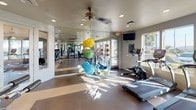 This fitness center is a thoughtfully designed, single-floor facility that offers a comprehensive workout environment for users of all fitness levels. The space is flooded with natural light through large windows that line the walls, providing open views of the surrounding outdoor greenery and creating a bright, energizing atmosphere for exercise.

The cardio zone is well-appointed with multiple True-brand treadmills, elliptical trainers, and a stationary bike, all arranged to take full advantage of the scenic window views. A wall-mounted flat-screen television adds to the overall experience. The flooring features a combination of durable rubber matting and smooth hardwood-toned sections, delineating workout zones effectively.

Strength training is equally well-served, with a multi-station cable machine and several selectorized weight machines from the True Fitness line positioned throughout the space. Floor-to-ceiling mirrors span one wall, amplifying the sense of space and allowing users to monitor their form during exercises.

A functional training area includes an assortment of colorful exercise balls, resistance bands, and other accessories neatly organized on a rack. A ceiling fan provides additional air circulation, complementing the recessed lighting overhead. The overall ambiance is clean, modern, and motivating, making this fitness center a fully capable facility for a well-rounded exercise routine.