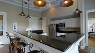 This single-floor space showcases a thoughtfully designed open kitchen that serves as its centerpiece. Rich, dark granite countertops extend across a generously sized breakfast bar, providing both a functional workspace and a casual dining area complete with ornate wrought iron bar seating. White raised-panel cabinetry lines the walls, offering ample storage while brightening the space and creating a clean contrast against the dark stone surfaces. Stainless steel appliances, including a full-size refrigerator and a built-in microwave, contribute a modern, polished aesthetic. Above, a coffered ceiling with decorative molding adds architectural depth and elegance to the room. A striking set of industrial-style pendant lights with wide, dome-shaped shades hang at varying heights over the bar area, creating a warm, ambient glow that complements the overall design. The neutral wall tones tie the space together, lending a versatile and inviting atmosphere. A doorway visible in the background suggests seamless flow to additional areas of the space. Overall, this kitchen blends classic detailing with contemporary finishes to create a functional and visually appealing environment.