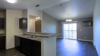 This single-floor space showcases an open-concept layout that seamlessly connects the kitchen and living areas, creating a bright and functional environment. The kitchen features dark cabinetry paired with granite-look countertops and a peninsula with a double sink, offering both practical workspace and a natural visual divider between the cooking and living zones. Pendant lighting above the peninsula adds a stylish focal point, complementing the overall design.

The living area benefits from vaulted ceilings that lend a sense of spaciousness and airiness to the space. A sliding glass door floods the room with natural light and provides direct access to a private balcony, offering outdoor views. An elegant chandelier hangs above the living area, adding a refined touch to the space.

The flooring throughout is a rich, dark hardwood-style material that ties the open-plan areas together cohesively. Neutral gray walls provide a clean, versatile backdrop that works well with both the dark cabinetry and the warm lighting fixtures. The overall ambiance is modern yet welcoming, with thoughtful design elements that balance functionality and comfort throughout the single-floor layout.