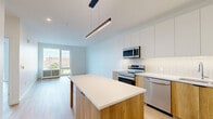 Located at 347 N Queen St in the heart of Lancaster, PA, this thoughtfully designed one-bedroom, one-bathroom unit offers a clean, contemporary aesthetic throughout. Light-toned hardwood flooring flows seamlessly across the open-plan living and kitchen areas, creating a cohesive and airy feel. The kitchen is a standout feature, boasting a two-tone design with crisp white upper cabinetry and warm wood-finish lower cabinets, complemented by a vertical tile backsplash, white quartz countertops, and a full suite of stainless steel appliances. A central island with an integrated dining extension provides additional workspace and casual seating. A sleek linear pendant light anchors the kitchen visually. The living area is bright and open, with large floor-to-ceiling windows offering sweeping views of the Lancaster cityscape and filling the space with abundant natural light. The bedroom is generously proportioned, featuring the same warm hardwood flooring, crisp white walls, expansive city-view windows, and a spacious sliding-door closet. The bathroom is finished with large-format neutral tiles, a glass-enclosed walk-in shower, and a wood-accented vanity with a clean white countertop, delivering a spa-like atmosphere.