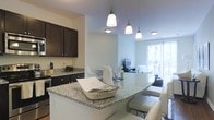 This single-floor residence offers a thoughtfully designed open-concept layout that seamlessly connects the kitchen, dining, and living areas into one cohesive and functional space. The kitchen is a standout feature, showcasing rich dark cabinetry that contrasts elegantly with speckled granite countertops. A curved kitchen island with bar seating serves as both a practical workspace and a social hub, illuminated by a trio of pendant lights that add a warm, inviting ambiance. Stainless steel appliances and ample cabinet storage complete the well-appointed kitchen. The dining area flows naturally from the kitchen, providing a comfortable space for everyday meals or entertaining guests. Beyond the dining area, the living space is bright and airy, benefiting from natural light that pours in through a large window or glass door that opens to an outdoor area, offering pleasant views and a seamless indoor-outdoor connection. The overall color palette of soft neutral walls, light flooring, and dark cabinetry creates a balanced and modern aesthetic throughout. The open plan design enhances the sense of spaciousness, making this residence feel welcoming and easy to navigate.
