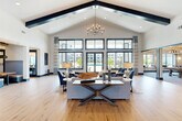 Located at 6735 Standifer Gap Rd in Chattanooga, Tennessee, this impressive single-story community clubhouse offers a generously scaled gathering space designed for both relaxation and social engagement. The architecture is defined by dramatic vaulted ceilings accented with bold dark exposed beams, creating a striking visual contrast against crisp white walls throughout the main lounge areas. Warm-toned wide-plank hardwood flooring flows through the primary seating zones, while elegant chandeliers serve as focal lighting fixtures that complement the upscale design aesthetic.

Floor-to-ceiling multi-pane windows flood the interior with abundant natural light and provide sweeping views of the outdoor pool area and surrounding landscaping, seamlessly connecting the interior to the exterior environment. Multiple distinct seating areas are thoughtfully arranged to accommodate a variety of social settings, from intimate conversation groupings to larger gatherings.

A dedicated game and entertainment room features tile flooring, a foosball table, casual dining tables, and a stylish wet bar area with a bold geometric tile backsplash. Additional areas include a conference-style workspace, private rooms accessible through dark-framed glass doors, and a large open reception area with shiplap wall detailing. The overall ambiance is modern yet inviting, blending contemporary design with comfortable, community-focused living.
