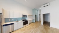 This single-floor unit features a contemporary design with a clean, open layout and consistent light hardwood flooring throughout. The main living area is notably spacious with soaring ceilings that amplify the sense of openness. A large sliding glass door floods the space with natural light while providing direct access to an outdoor area. A two-tone wall treatment — white above and warm beige below — adds subtle visual interest to the living space.

The kitchen is well-appointed with a full suite of stainless steel appliances, including a refrigerator, electric range, built-in microwave, and dishwasher. White flat-front cabinetry pairs with white quartz countertops and a soft blue tile backsplash, creating a bright, modern aesthetic. Open wood floating shelves add a warm, functional accent. A wall-mounted mini-split HVAC unit is also present.

Additional rooms include a private bedroom with bright white walls and hardwood floors, as well as a full bathroom featuring decorative patterned floor tiles in gold and grey, a tiled tub-shower combination, and a white vanity. The unit also includes a dedicated closet or storage area, as visible in the floor plan view.