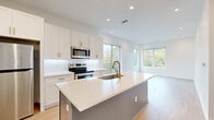 This contemporary single-floor space showcases a clean, modern aesthetic defined by bright white walls, light hardwood-style flooring, and an abundance of natural light. The open-concept layout seamlessly connects the kitchen and living area, creating a generous and airy feel throughout. Large windows frame lush, tree-lined views and allow sunlight to pour in from multiple directions, enhancing the overall sense of openness.

The kitchen is a standout feature, centered around a substantial island with crisp white quartz countertops and an integrated undermount sink topped with a brushed gold faucet. White shaker-style cabinetry with gold hardware lines the walls, complemented by a clean subway tile backsplash and a full suite of stainless steel appliances, including a range, over-the-range microwave, and refrigerator. Recessed lighting throughout provides consistent, well-distributed illumination.

The bedroom offers plush carpeted flooring, recessed lighting, and generous window placement with serene outdoor views. The bathroom is well-appointed with a white shaker vanity, quartz countertop, gold hardware accents, and a large frameless mirror. A private balcony accessible from the living and kitchen area further extends the living space outdoors, adding an inviting connection to the surrounding natural landscape.