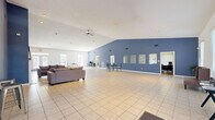 Located at 100 Remington Dr in Dickson, Tennessee, this single-story community clubhouse offers a generously sized, open-plan interior designed to serve a variety of social and recreational functions. The main gathering area is defined by soaring vaulted ceilings and a ceiling fan, creating an airy, welcoming atmosphere throughout. Light-toned ceramic tile flooring extends across the entire space, complementing the crisp white ceilings and bold blue accent walls that lend the interior a clean, modern character. A cozy lounge zone anchors one end of the main hall, complete with a white fireplace mantel and comfortable seating arrangements that invite relaxed conversation. Large windows along the perimeter allow natural light to fill the open floor plan. A kitchenette area is equipped with a bar-height counter, bar stools, cabinetry, and a full-size refrigerator, making it well-suited for community gatherings and events. A dedicated fitness center, accessible via a glass-panel door, provides a separate space for exercise equipment. Along one wall, a row of pass-through windows opens to adjacent office or administrative spaces, supporting the facility's functional needs. The main entrance features French doors with sidelight windows, creating a bright and inviting entryway. Overall, the space is thoughtfully designed to accommodate community events, fitness, and everyday amenities in a comfortable, well-lit environment.