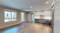 North Grove A1D is a thoughtfully designed 720-square-foot, one-bedroom, one-bathroom apartment located in Riverside, CA 92501. The single-floor layout includes a kitchen, bedroom, bathroom, hallway, and private balcony, offering an efficient and comfortable living arrangement. The kitchen is a standout feature, showcasing grey shaker-style cabinetry, a white subway tile backsplash, quartz countertops, and a full suite of stainless steel appliances including a French door refrigerator, range, microwave, and dishwasher — all set atop light wood-look flooring. The 120-square-foot bedroom is finished with soft wall-to-wall carpet, neutral grey walls, and a large window that allows natural light and views of the modern surrounding complex. The bathroom features light wood-look flooring, a combined tub and shower, a toilet, and a vanity with blue-grey shaker cabinetry topped with a dark countertop and a large mirror. The private balcony overlooks the community's contemporary multi-story buildings and parking area under sunny Southern California skies. The overall aesthetic is clean and modern, with a neutral color palette, recessed lighting, and consistent finishes throughout that give the space a fresh, move-in-ready feel.