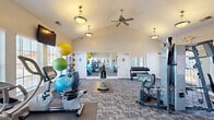 This fitness center is thoughtfully designed to support a full range of workout routines within a spacious, single-floor layout. The architectural highlight is the soaring vaulted ceiling, which lends an open, airy atmosphere throughout the main exercise area. Warm beige walls, patterned carpet tile flooring, and elegant pendant lighting create a welcoming environment, while a ceiling fan promotes comfortable airflow during workouts.

The cardio zone is well-appointed with multiple Matrix and True treadmills, ellipticals, an Octane elliptical, and recumbent and upright stationary bikes, all positioned along large windows fitted with horizontal blinds that allow ample natural light. Wall-mounted televisions provide entertainment during cardio sessions.

The strength and functional training area features a cable resistance machine, a tiered dumbbell rack, resistance bands, and an assortment of stability and medicine balls in various sizes. Large floor-to-ceiling mirrors flank the rear wall, visually expanding the space and aiding in form correction.

Two restrooms are conveniently accessible from the main floor. The overall ambiance is clean, bright, and functional, offering a well-rounded fitness environment suitable for a variety of exercise preferences.