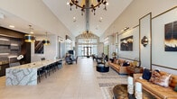 Located at 16230 N Litchfield Rd in Surprise, Arizona, this elegantly designed single-floor clubhouse offers a generously proportioned layout tailored for both social gatherings and productive work. The centerpiece is a dramatic open-plan kitchen and living area boasting soaring vaulted ceilings adorned with striking chandelier fixtures, large-format light tile flooring, and an abundance of recessed lighting. The living area features stylish leather seating, bold contemporary artwork, and a sleek paneled accent wall with an integrated flat-screen display, creating an inviting yet refined atmosphere. Adjacent to the living space, the dining area presents a grand marble-topped table beneath paired gold pendant lights set against a dark accent wall, providing a sophisticated setting for group dining or meetings. A dedicated hallway connects additional flexible spaces, including a conference-style room with a solid wood table, built-in workstations, and tall windows that draw in natural light and outdoor views. A game room with a shuffleboard table and large picture windows further adds to the recreational appeal. Throughout, the space maintains a cohesive modern aesthetic with carefully curated décor, textured wall treatments, and premium finishes that collectively establish an upscale, community-focused environment.