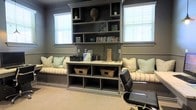 This single-floor space is thoughtfully designed to blend functionality with comfort. The focal point of the room is a floor-to-ceiling built-in shelving unit finished in a muted gray-green tone, flanked by two windows fitted with white horizontal blinds that allow ample natural light to filter in. The shelving provides generous display and storage space, housing books, decorative objects, and organizational accessories.

Beneath the upper shelving, a built-in workspace features a light-toned surface ideal for a home office or study area. Below the desk level, open cubbies with woven baskets offer additional organized storage. Flanking the central shelving unit on both sides, built-in bench seating runs the length of the wall, upholstered in a neutral stripe fabric and accented with soft sage green and textured throw pillows, creating a comfortable lounge area.

The walls are finished in a coordinating warm gray tone and feature classic wainscoting detail, adding architectural interest. Neutral carpeting grounds the space, while a black leather office chair completes the practical workspace setup. The overall ambiance is polished, organized, and welcoming.