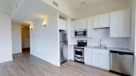 This single-floor 1-bedroom, 1-bathroom residence offers a thoughtfully designed layout with a contemporary aesthetic. The kitchen is a standout feature, showcasing white shaker-style cabinetry that extends to the ceiling, maximizing storage capacity. Stainless steel appliances include a full-size refrigerator, a range with oven, and an over-the-range microwave, all complementing the space's modern character. Light granite countertops and a classic white subway tile backsplash add texture and visual interest, while a single-basin sink with a modern faucet completes the functional workspace. The kitchen flows naturally into the adjacent living area, creating an open, connected feel. Light-toned hardwood-style flooring runs throughout, lending warmth and continuity to the space. Soft wall sconces provide ambient lighting, enhancing the clean, neutral color palette of white walls and light trim. Ceiling vents suggest a central HVAC system for year-round comfort. The overall ambiance is clean, modern, and move-in ready, appealing to those seeking a well-appointed, low-maintenance living space.