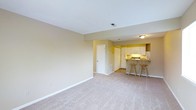 This single-floor space presents an open-concept layout that connects a spacious living area to a functional kitchen. The living area is finished with wall-to-wall carpeting in a neutral tone, complemented by light beige walls and white baseboards that contribute to a clean, understated aesthetic. Ceiling-height is standard, with a flush-mounted light fixture providing overhead illumination.

The kitchen is positioned at the rear of the space and features white cabinetry both above and below the counters, offering ample storage. A raised breakfast bar separates the kitchen from the living area and is accompanied by two wooden bar stools, creating a casual dining option. The kitchen is equipped with a range and a full-size refrigerator. Additional built-in storage or closet space is accessible via a white panel door adjacent to the kitchen area.

A floor vent is visible near the kitchen transition, indicating a forced-air climate system. The overall layout is efficient and straightforward, making practical use of the available space while maintaining an open, cohesive feel throughout the main living and kitchen zones.