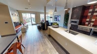 This single-floor space is designed with an open-plan layout that seamlessly connects a kitchenette area to a comfortable lounge. The kitchenette features a long, light-colored countertop with an integrated stainless steel sink, complemented by dark wood cabinetry and built-in stainless steel appliances, including a wall oven and microwave. Warm wood-look flooring extends throughout the entire space, contributing to a welcoming and cohesive aesthetic. Pendant lighting in neutral tones hangs above the countertop, while recessed ceiling lights provide additional illumination throughout. The lounge area is furnished with dark upholstered seating arranged around a central area, with a mounted flat-screen display on the wall. Lush potted plants are strategically placed to add a natural, vibrant element to the interior. At the far end of the space, large glass doors flood the interior with abundant natural light and offer a direct view and access to an outdoor courtyard, where palm trees and outdoor seating are visible. The overall ambiance is modern, clean, and inviting, making it well-suited for gathering and social interaction.