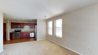 This single-floor space offers an open and functional layout that seamlessly connects the living area to the kitchen. The living area is finished with plush, neutral-toned carpeting that adds warmth underfoot, while two windows fitted with horizontal blinds allow natural light to brighten the space and provide views of the surrounding exterior. The clean, light beige walls and white ceiling contribute to an airy, understated aesthetic throughout.

The kitchen is positioned adjacent to the living area and features rich, reddish-brown cabinetry that provides ample storage. Stainless steel appliances, including a refrigerator and dishwasher, lend a modern touch, while a sink with under-cabinet lighting adds practicality. The kitchen is finished with hard-surface flooring, creating a clear visual distinction from the carpeted living space. A ceiling-mounted recessed light provides focused illumination over the kitchen area, complementing the natural light from the windows. Overall, the space presents a clean, move-in-ready environment with a balance of functional design and comfortable living features.