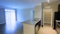 This single-floor space is designed with an open-concept layout that promotes a sense of airiness and functional flow throughout. The kitchen serves as a central hub, anchored by a large island with granite countertops, an undermount sink, and sleek dark cabinetry paired with stainless steel appliances. Light wood-tone flooring in the kitchen transitions smoothly into the adjoining living area, which features neutral-toned carpeting and soft, light-gray walls that create a clean and calming atmosphere. Vertical blinds along a large window allow for adjustable natural light, brightening the expansive living space. A doorway from the main living area leads into an additional room, offering flexible use of the layout. To the side, a walk-in closet with built-in shelving provides generous storage capacity. The overall design is contemporary and understated, with consistent neutral tones, clean lines, and practical finishes throughout. The open sightlines between the kitchen and living areas make the space feel connected and well-proportioned, well-suited for both everyday living and entertaining.