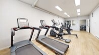Located at 134 College Hwy in Southampton, Massachusetts, this combined gym and laundry facility offers a practical and well-organized space across a single floor. The fitness area is bright and clean, featuring light wood-style flooring and crisp white walls that create an open, energizing atmosphere. Recessed panel lighting illuminates the space evenly, while windows allow natural light to filter in. The gym is outfitted with a variety of cardio and strength equipment, including treadmills, an elliptical trainer, a recumbent exercise bike, a multi-function cable machine, and a two-tier dumbbell rack stocked with a full range of free weights. Baseboard heating ensures year-round comfort. The laundry room is equally functional, housing a row of front-loading LG washing machines finished in sleek metallic silver, complemented by matching dryers. White louvered cabinetry mounted above the machines provides organized storage. The same light wood flooring carries through this area, maintaining a consistent, tidy aesthetic throughout. An entry area with a classic arched-window door provides access to the space. Together, these two areas form a convenient and well-maintained amenity facility suited for residential or community use.