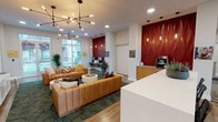This single-floor leasing lobby for Flora is designed to make a strong first impression through a cohesive blend of bold aesthetics and functional amenities. The entry corridor features large floor-to-ceiling windows that flood the space with natural light and provide views of the exterior grounds, with warm wood-look plank flooring running throughout.

The central lounge area anchors the space with a pair of tufted caramel leather sofas arranged around a pair of round white coffee tables, all set atop a lush botanical-print area rug. A striking sculptural sputnik-style chandelier hangs overhead, complementing the warm Edison-style pendant lights positioned throughout. Tall indoor palms and tropical plants add organic texture and reinforce the nature-inspired branding.

A dedicated business center lines one wall beneath a dramatic deep-red, three-dimensional wave-patterned accent wall, housing a row of computer workstations beneath pendant lighting — ideal for residents' daily productivity needs.

Adjacent to the lounge, a sleek white bar-height counter with wire mesh barstools serves as a gathering spot, flanked by warm wood cabinetry, a beverage refrigerator, and a coffee machine station — all set against another bold red accent wall. The overall ambiance is contemporary, vibrant, and welcoming, reflecting a community-centered design philosophy.