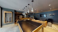 Located at 572 21st St in Oakland, CA, this single-floor entertainment space is thoughtfully designed to deliver an immersive, upscale lounge experience. The interior is defined by deep teal and dark charcoal walls that establish a bold, moody atmosphere throughout. Polished concrete flooring contrasts with a geometric patterned area rug, anchoring the central game area where a full-size ping-pong table and a solid wood pool table take center stage. A series of white pendant lights hang in a linear arrangement overhead, casting focused illumination across the game zone. A tufted brown leather sofa lines one wall, accompanied by a set of oval porthole-style mirrors that add a nautical-inspired design touch. The lounge area features a caramel-toned modular leather sofa, textured throw pillows, and a sculptural side stool, creating a relaxed gathering space. A wall-mounted flat-screen television and sleek wall sconces define the media corner, while a long wooden console provides display and storage. Natural light filters in through a glass door leading to an outdoor area. The space is further enhanced by an in-wall split AC unit, recessed ceiling speakers, and curated wall art, making it a well-equipped venue for both recreation and relaxation.
