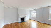 This single-floor space thoughtfully combines classic architectural character with contemporary updates throughout. The expansive living area serves as the centerpiece, showcasing warm hardwood floors, crown molding, and an ornate white marble fireplace mantel with intricate carved detailing. Multiple large windows line the walls, flooding the room with natural light and offering views of the surrounding neighborhood streetscape.

The bedroom areas also feature the same rich hardwood flooring and crown molding, maintaining a cohesive aesthetic. Each room benefits from generous natural light through classically trimmed windows, and pendant light fixtures provide additional illumination.

The kitchen has been fully updated with a clean, modern sensibility. White shaker-style cabinetry with glass-front uppers lines the walls, complemented by a marble-style tile backsplash, dark stone countertops, and stainless steel appliances including a gas range, microwave, and dishwasher. A central island with a dark countertop adds prep space, while globe pendant lights hang overhead. Large transom windows bring in additional natural light.

The bathroom is finished in neutral tones with large-format tile walls and floors. It features a dark-cabineted vanity with a white countertop, a framed mirror, wall sconces, and a glass-enclosed walk-in shower, creating a clean and functional space.