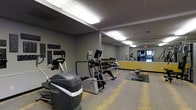 The Timbers fitness center is a well-equipped, single-floor exercise space designed to support a variety of workout routines. The room features a selection of cardio machines, including ellipticals and a stair climber, arranged to allow comfortable use and movement throughout the space. A dumbbell rack lines one wall, offering a range of free weights for strength training. A large floor-to-ceiling mirror spans one wall, visually expanding the room and allowing users to monitor their form during workouts. The flooring consists of a combination of hard surface and rubber exercise mats, providing both durability and cushioning where needed. Overhead fluorescent lighting ensures the space is well-lit throughout. The walls are painted in a contemporary gray palette accented with yellow trim, giving the room a modern, energetic feel. Motivational signage featuring words such as "Achieve," "Conquer," and "Dedication" adds an encouraging atmosphere. The textured ceiling and structural column are consistent with the building's practical design. Overall, the fitness center offers a functional and motivating environment for residents looking to maintain an active lifestyle.