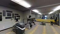 The Timbers fitness center is a well-equipped, single-floor exercise space designed to support a variety of workout routines. The room features a selection of cardio machines, including ellipticals and a stair climber, arranged to allow comfortable use and movement throughout the space. A dumbbell rack lines one wall, offering a range of free weights for strength training. A large floor-to-ceiling mirror spans one wall, visually expanding the room and allowing users to monitor their form during workouts. The flooring consists of a combination of hard surface and rubber exercise mats, providing both durability and cushioning where needed. Overhead fluorescent lighting ensures the space is well-lit throughout. The walls are painted in a contemporary gray palette accented with yellow trim, giving the room a modern, energetic feel. Motivational signage featuring words such as "Achieve," "Conquer," and "Dedication" adds an encouraging atmosphere. The textured ceiling and structural column are consistent with the building's practical design. Overall, the fitness center offers a functional and motivating environment for residents looking to maintain an active lifestyle.
