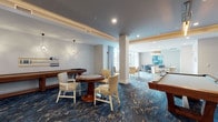 Located at 265 Communication Way in Hyannis, Massachusetts, this single-floor amenity space offers a thoughtfully designed social hub with distinct activity zones seamlessly connected by rich, ocean-inspired blue patterned carpeting throughout. The game room area is a standout feature, equipped with a shuffleboard table, a poker/card table with patterned upholstered chairs, and a billiards table, all accented by decorative oar wall art and elegant pendant lighting that evoke a nautical Cape Cod aesthetic. Adjacent to the game room, a comfortable lounge area features modern tufted armchairs arranged around a woven coffee table, anchored by a textured stone-panel accent wall displaying multiple flat-screen televisions. A tray ceiling with warm cove lighting adds architectural depth to this relaxation zone. The dining and gathering area is bathed in natural light from expansive floor-to-ceiling windows and glass doors that open directly onto an outdoor terrace, offering views of the surrounding greenery. Multiple dining tables with eclectic patterned chairs create a welcoming space for group activities, including a dedicated puzzle and game table positioned beside the terrace doors. The overall design blends coastal charm with modern comfort, creating an inviting, multi-functional space ideal for socializing and recreation.