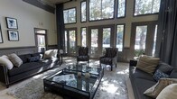 This refined single-floor space makes an immediate impression with its dramatic double-height ceilings and a sweeping wall of large, dark-framed windows that flood the interior with abundant natural light. The floor-to-ceiling glazing offers picturesque views of lush outdoor greenery, seamlessly connecting the interior to the surrounding landscape.

The lounge area is thoughtfully appointed with a cohesive palette of deep charcoal and grey tones. Plush, patterned wingback chairs are arranged around a sleek glass-top coffee table, complemented by a pair of substantial sofas and a soft, textured area rug that anchors the seating arrangement. The glass table surfaces reflect the natural light, enhancing the spacious and airy feel of the room.

Tall, elegant drapery panels flank the windows, adding a layer of sophistication and texture to the space. The neutral wall tones provide a refined backdrop that allows the furnishings and architectural details to stand out. The combination of soaring ceilings, generous fenestration, and carefully curated furnishings gives this space a grand yet comfortable ambiance, making it well-suited for both relaxed gatherings and more formal social interactions.